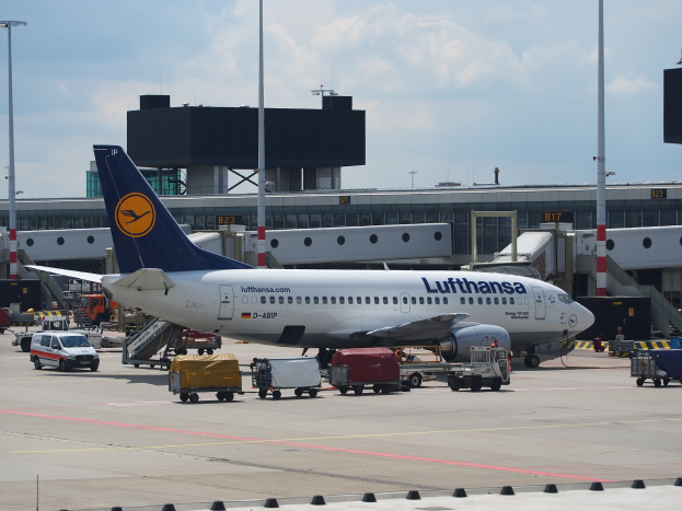 Ein Lufthansa Airbus A320-200-Flugzeug am Frankfurter Flughafen mit Bodenfahrzeugen und Wagen im Vordergrund, einem Geb"ude und Laternen im Hintergrund und einem klaren blauen Himmel.