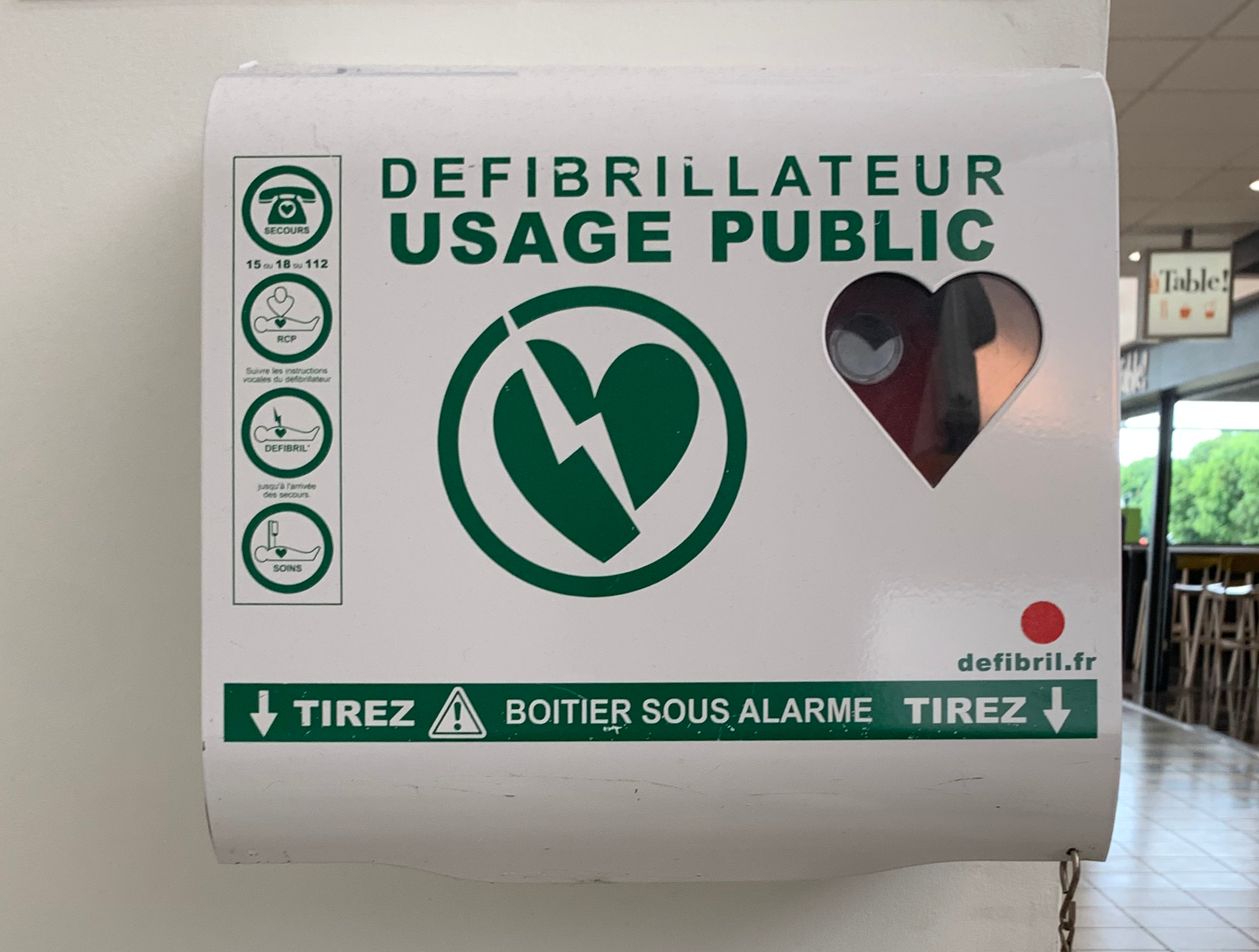 Ein öffentliches Defibrillator-Gebrauchszeichen an einer Gebäudewand, mit Stühlen, Tischen, Plakaten, Lampen und Bäumen im Hintergrund und dem Boden unten sichtbar.
