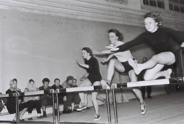 Zwei Frauen in der Luft über ein Hürdenhindernis springend in einer Turnhalle, mit Zuschauern links und Fenstern an der Wand im Hintergrund.