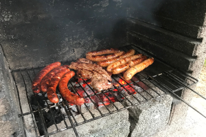 Grill mit Würstchen und Fleisch darauf, umgeben von brennender Kohle und Flammen, im Freien auf Gras.