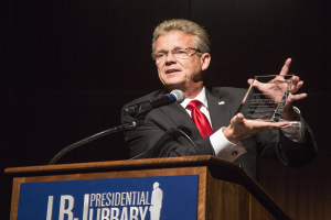 Ein Mann in Anzug und Krawatte, der eine Auszeichnung auf einem Podium mit Mikrofon und einer Tafel hält, vor einem dunklen Hintergrund bei einer Bibliothek-Veranstaltung.