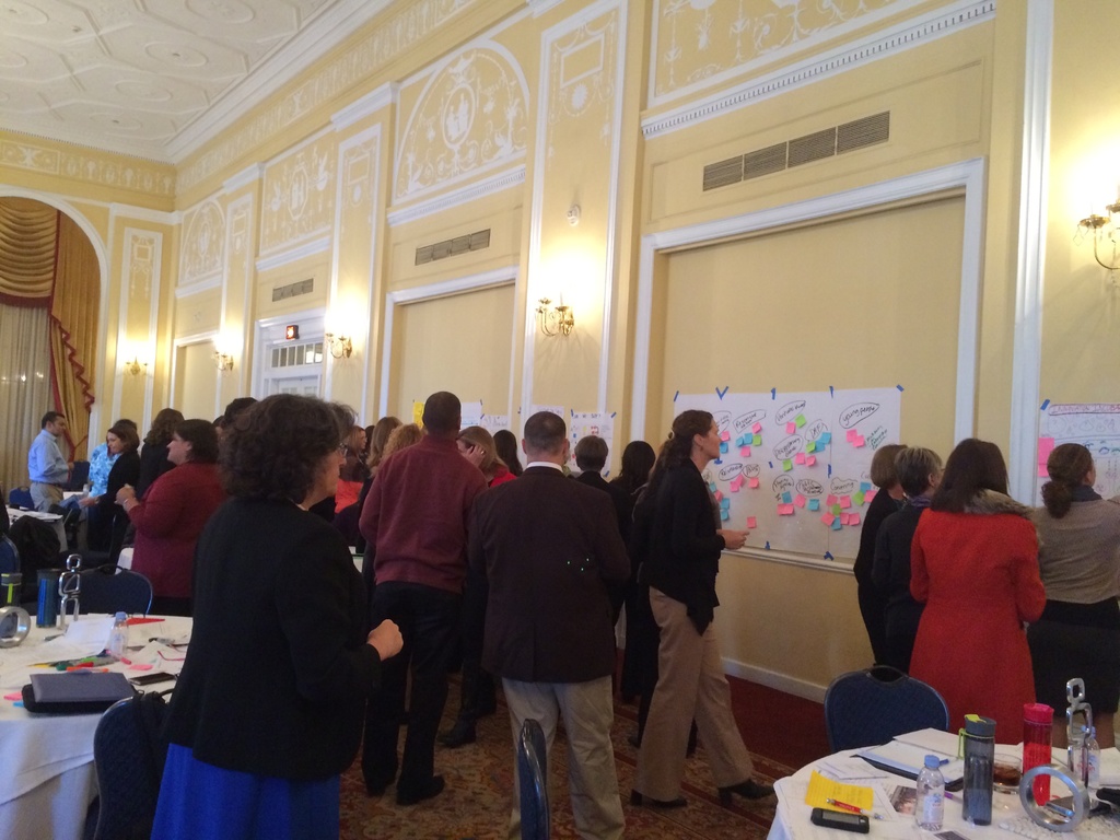 Eine Gruppe von Menschen untersucht Plakate und Diagramme, die an einer Wand in einem Raum zu sein scheinen, der wie ein Kunstmuseum aussieht.