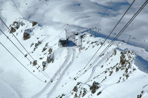 Eine Luftaufnahme eines Skilifts, der einen schneebedeckten Berg hinabfährt, mit sichtbaren Seilbahnkabeln, Felsen und Häusern, bei strahlendem Sonnenschein.