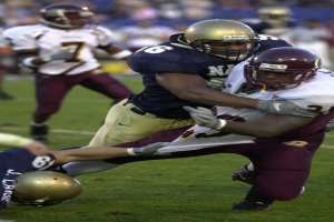 Zwei Fußballspieler in Helmen und Uniformen stoßen auf dem Feld zusammen, während andere im Hintergrund zuschauen.
