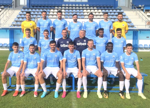 Eine Gruppe von Männern, einige stehen und einige sitzen auf Bänken, auf einem Fußballfeld, mit Stühlen, Treppen, Geländern, Wänden und Fenstern im Hintergrund.