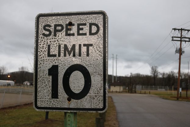 Geschwindigkeitsbegrenzungstafel am Straßenrand mit Gras, Strommästen, einem Zaun, Häusern, Bäumen und einem bewölkten Himmel im Hintergrund.