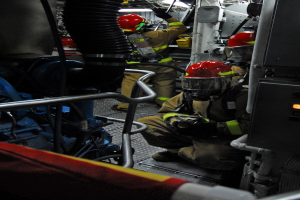 Feuerwehrleute in Schutzausrüstung arbeiten an einem Löschwagen mit Equipment im Hintergrund und einer Lampe oben.