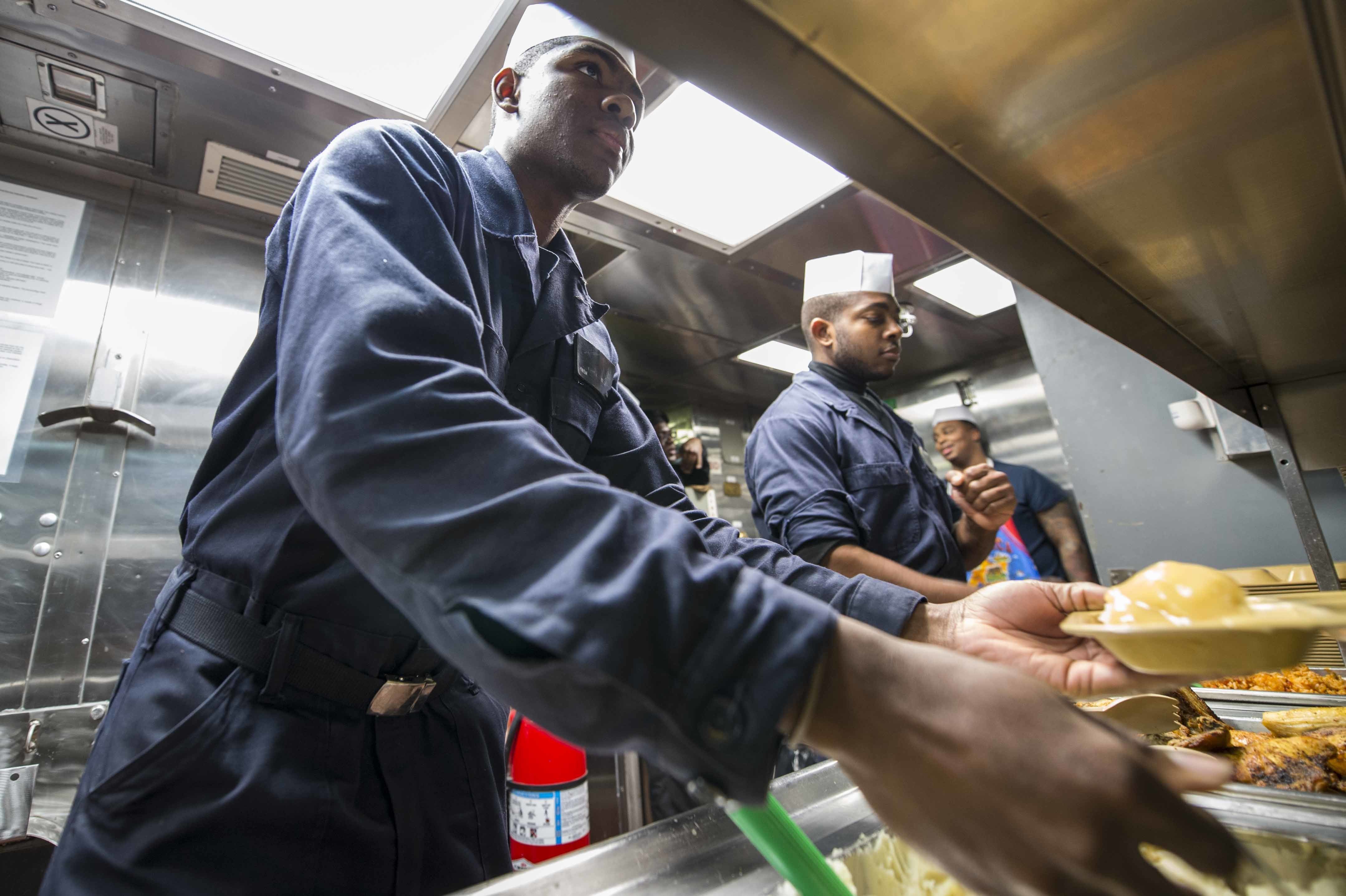 Eine Gruppe Von M├Ąnnern Steht In Einer K├╝che, Einer Hält Ein Teller Mit Essen, Mit Tabletts Von Essen Auf Dem Tisch, Einem Feuerl├Âscher Und Papieren An Der Wand Im Hintergrund.