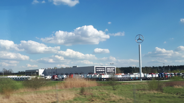 Ein Mercedes-Benz-Werk in Deutschland mit Fahrzeugen auf der Straße, umgeben von Gras, Pflanzen, Bäumen, Gebäuden und Polen, von denen einer ein Logo trägt.